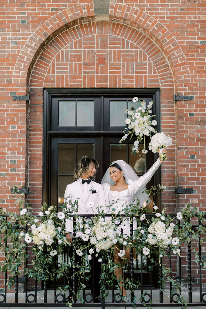 Couple in tux and wedding gown standing on a balcony with female holding bouquet of flowers in the air.